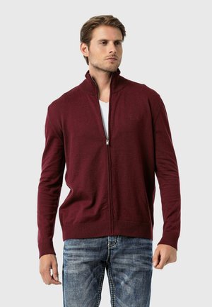 Burgunderfarbener Reißverschluss-Pullover mit hohem Kragen, gerippten Bündchen und einem strukturierten Strickdesign. Getragen über einem weißen T-Shirt mit blue Jeans.