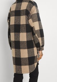 Lång rutig kappa i beige och svart. Tillverkad av textil med struktur och rundad fåll, med överdimensionerade rutmönster och uppvikta ärmar.