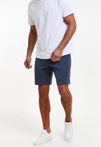 Navy šortky se strukturovaným povrchem, s páskem na stažení v pase a malou nášivkou s logem. V kombinaci s jednoduchým bílým tričkem a bílými teniskami.