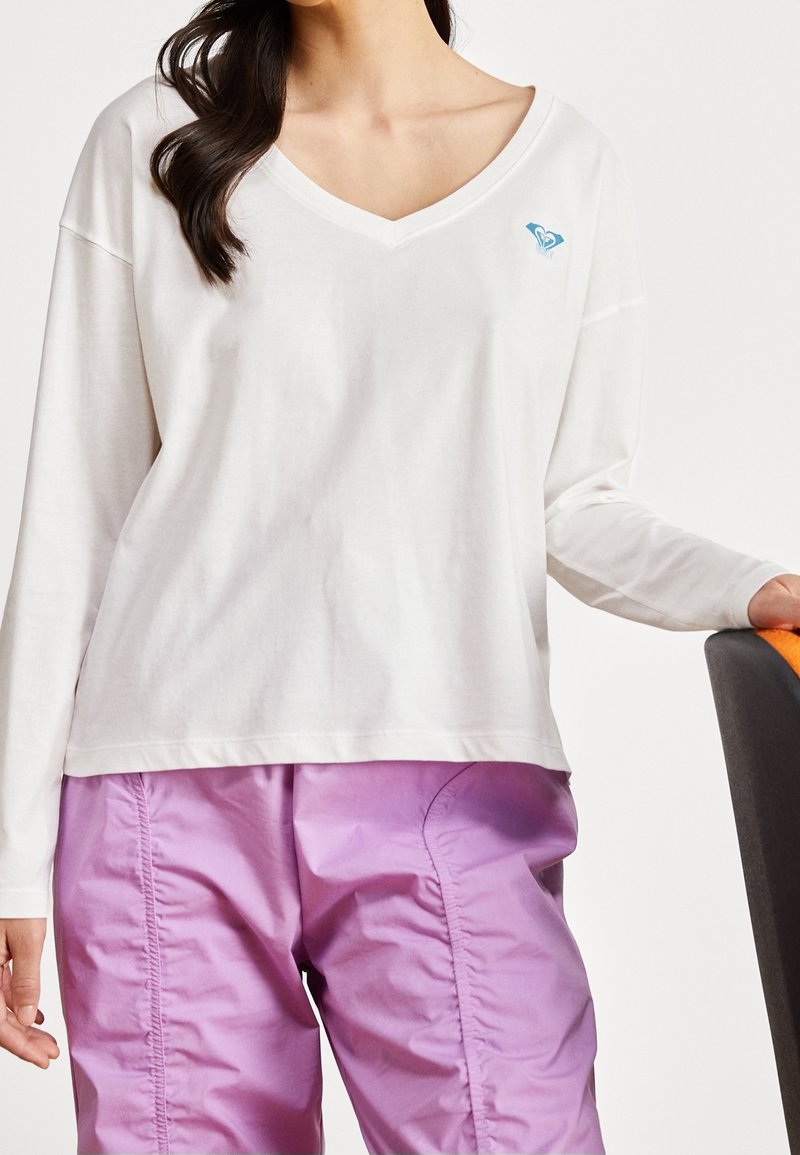 Femme portant une chemise blanche à col en V à manches longues avec un petit logo bleu et un pantalon violet brillant, reposant une main sur une chaise.