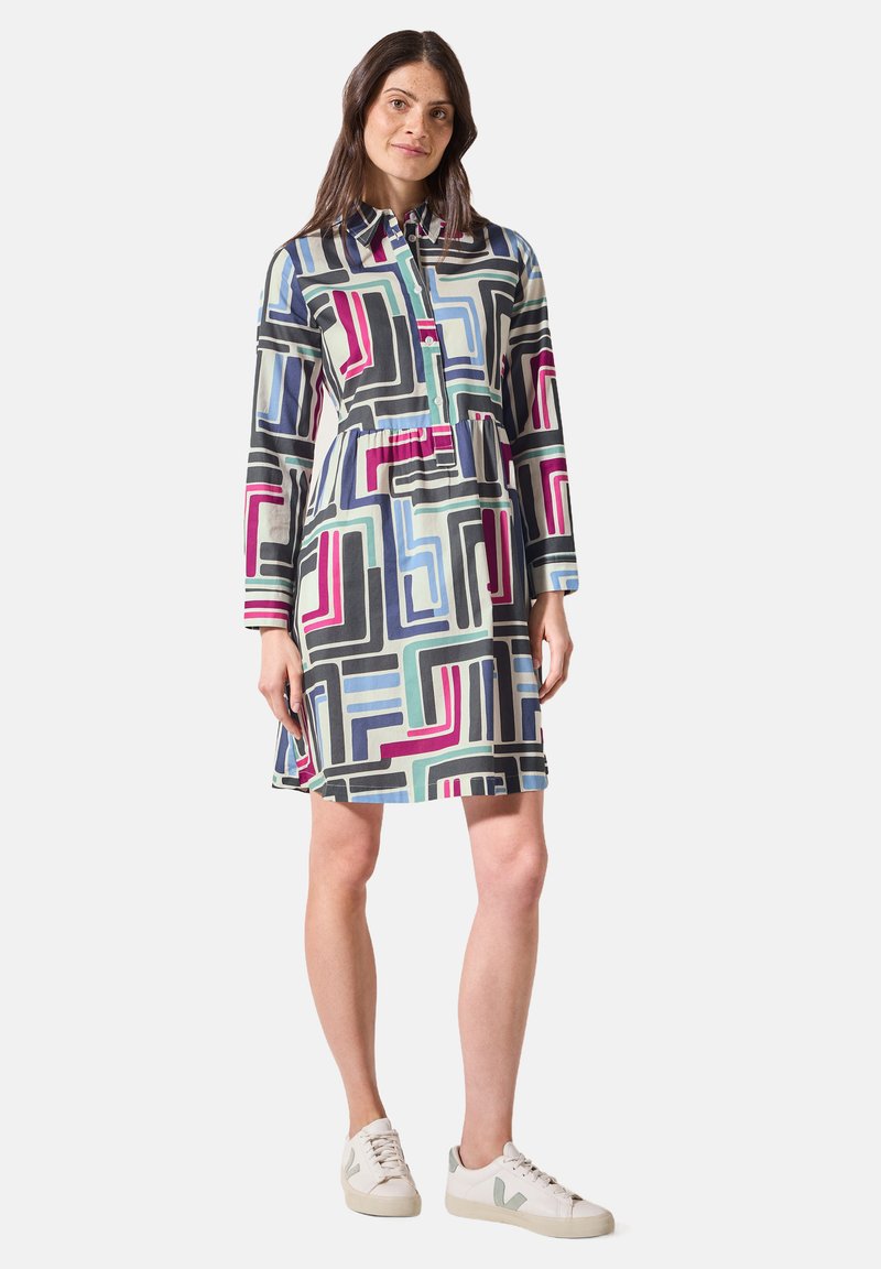 Button-up Hemdkleid im mehrfarbigen geometrischen Muster. Hat lange Ärmel, eine taillierte Taille und einen geraden Schnitt. Getragen mit weißen Turnschuhen.