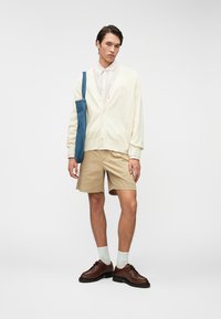 Νεαρός άνδρας με μπεζ σορτς, κρεμ κασκόλ, ριγέ πουκάμισο, καφέ παπούτσια, λευκές κάλτσες και κρατώντας μια μπλε τσάντα ώμου, που στέκεται σε λευκό φόντο.