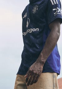 Fioletowa koszulka Manchester United z teksturowanym wzorem, białymi akcentami oraz trzema pionowymi paskami na każdym ramieniu.