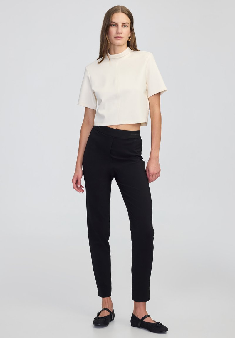 Haut créme court avec des manches courtes et un col montant, associé à un pantalon noir ajusté et des chaussures plates noires. Tissu lisse, design minimalist.