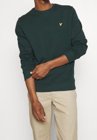 Sudadera de cuello redondo de color verde oscuro, hecha de algodón, con puños y dobladillo acanalados, y un pequeño logotipo amarillo en el lado izquierdo del pecho.