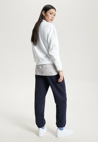 Felpa bianca con polsini a costine, indossata sopra una maglietta grigia chiara con motivi floreali rossi, abbinata a joggers blu navy e sneaker bianche.