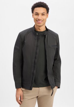 Glunderende man met een zwart jack met rits over een donker shirt en beige broek, staand met één hand in de zak tegen een witte achtergrond.