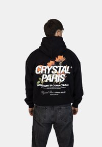 Černá mikina s kapucí s velkým bílým nápisem "CRYSTAL PARIS" a květinovými grafikami. S volným střihem a žebrovanými manžetami. Nosená s tmavými džíny.
