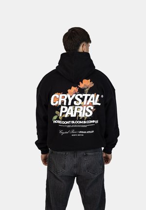 Black hoodie featuring large white text "CRYSTAL PARIS" and floral graphics. Hooded with a relaxed fit and ribbed cuffs. Worn with dark denim.