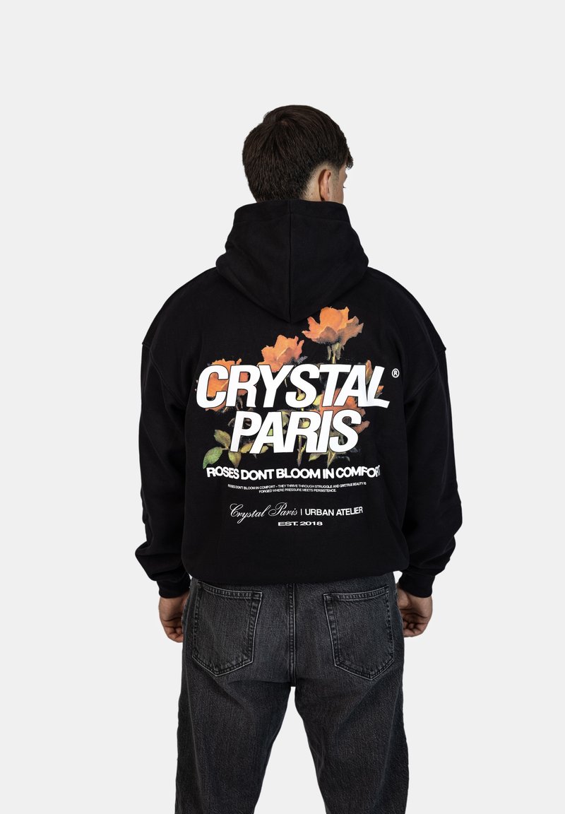 Černá mikina s kapucí s velkým bílým nápisem "CRYSTAL PARIS" a květinovými grafikami. S volným střihem a žebrovanými manžetami. Nosená s tmavými džíny.