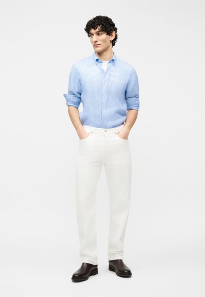 Άνδρας με σγουρά μαλλιά που φοράει ανοιχτό μπλε ριγέ πουκάμισο, άσπρο παντελόνι και σκούρα καφέ μπότες, στέκεται με τα χέρια στις τσέπες.