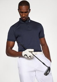 Polo azul marinho com colarinho, mangas curtas e logótipo no colarinho; modelo a usar calças brancas e a segurar uma taco de golfe com uma luva.