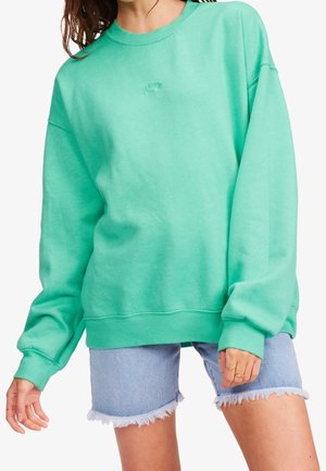 Donna che indossa un ampio sweatshirt verde menta e pantaloni shorts in denim chiaro strappati, mostrando anelli sulle sue dita.