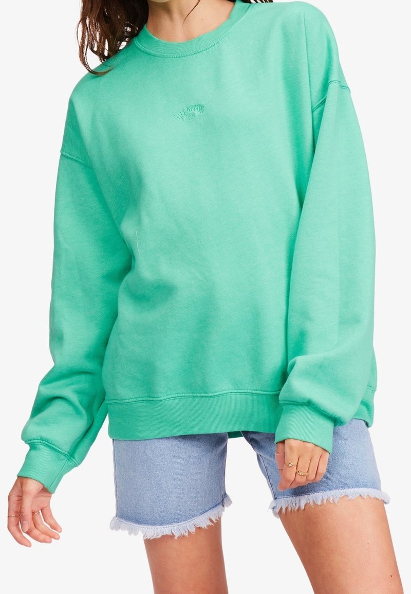 Kvinna som bär en överdimensionerad mintgrön sweatshirt och ljusblå fransiga denimshorts, som visar ringar på sina fingrar.