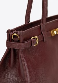 Bordowa skórzana torebka z złotymi akcentami, o strukturalnym kształcie i teksturowanej powierzchni. Posiada podwójne uchwyty i zabezpieczone zamknięcie na górze.