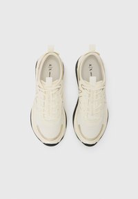 Cremefarbene Sportschuhe mit Obermaterial aus Textil, schwarzer Sohle, goldenen Akzenten und Schnürsenkeln; verfügen über eine Zuglasche und ein markiertes Innenetikett.