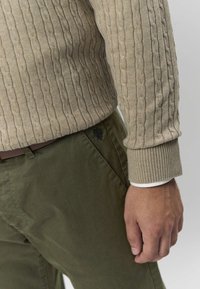 Beżowy sweter z dzianiny z wzorem warkocza, zestawiony z oliwkowozielonymi spodniami z subtelnym haftowanym detalem. Kontrast tekstur w tkaninie.