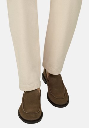 Braune Wildleder-Slipper, getragen mit beigen Hosen, im Fokus die Unterschenkel und Füße vor neutralem Hintergrund.