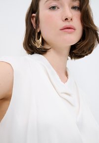 Top bianco senza maniche con scollo drappeggiato e texture morbida e liscia; modella che indossa grandi orecchini d'oro circolari.