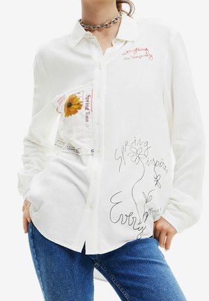 Kobieta ubrana w białą koszulę na guziki z kwiatowymi i tekstowymi wzorami, w połączeniu z niebieskimi dżinsami i srebrnym naszyjnikiem z łańcuszkiem.