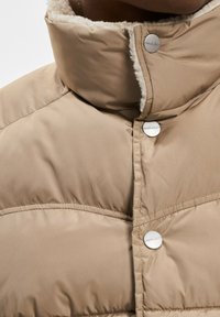 Beige polstret jakke med høj krave, blød indvendig foret, og sølv knapper. Glat ydre med mat finish og vandrette syninger.