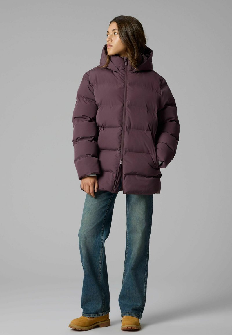 Femme portant une doudoune violette avec capuche, jean bleu et bottes couleur cuir clair, debout et regardant vers sa gauche sur un fond gris.