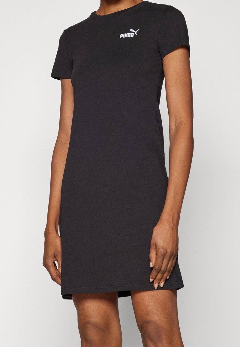 Personne portant une robe noire Puma à manches courtes, longueur genou, avec un logo blanc sur la poitrine, debout devant un fond uni.