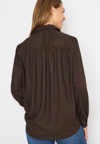 Braune Bluse mit langen Ärmeln, einem Rückenschlitz, weichem Stoff, geknöpften Manschetten und einem klassischen Kragen, kombiniert mit blauen Jeans.