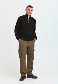 Schwarzer Strickpullover mit Viertelreißverschluss, kombiniert mit olivfarbenen Cargo-Hosen, die mehrere Taschen haben, sowie schwarzen Schuhen mit strukturierter Optik.