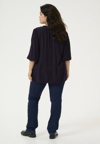 Blusa púrpura oscura con mangas cortas abullonadas, corte suelto, escote redondo, combinada con jeans azul oscuro y zapatos negros. Diseño sencillo, sin patrones.