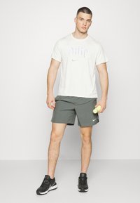 Grå sportshorts, vit T-shirt med tryckt logotyp, svarta sneakers, håller i en grön hantel. En enkel design med funktionella träningskläder.