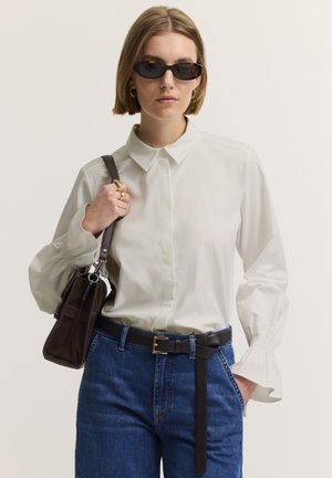 Frau trägt weiße Bluse, blaue Jeans, schwarzen Gürtel, dunkle Sonnenbrille, goldene Creolen, hält eine braune Handtasche auf der Schulter.