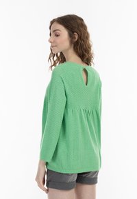 Top verde con texture, stile peplum, maniche a tre quarti e dettaglio a chiave sul retro. Abbinato a pantaloni corti grigi.