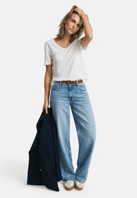 Weißes T-Shirt mit kurzen Ärmeln, hellblaue weit geschnittene Jeans, brauner Gürtel, hält ein marineblaues Sakko, legere Sneakers, minimalistisches Design, lockere Passform.