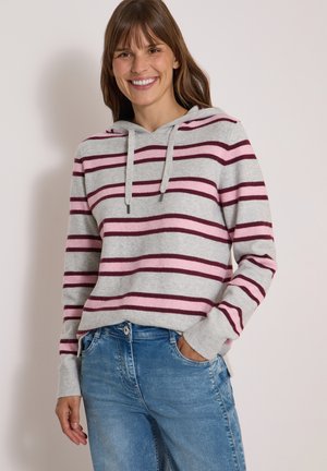 Femme souriante portant un sweat à capuche gris avec des rayures roses et bordeaux, associée à un jean bleu, debout devant un fond clair uni.