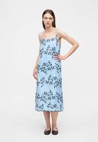 Γυναίκα όρθια με το χέρι στη μέση, φορώντας γαλάζιο φόρεμα μίντι με μαύρα λουλουδάτα κεντήματα και κυματιστό στρίφωμα, συνδυασμένο με σκούρα παπούτσια χωρίς τακούνι.