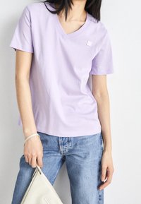 Lila bomulls T-shirt med V-hals och korta ärmar, tillsammans med ljusblå jeans och en krämfärgad väska med texturerad logodetalj.