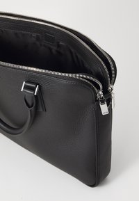 Sacoche pour ordinateur portable en cuir noir avec une finition texturée, fermetures à double zip et une poignée robuste. L'intérieur est doté d'une doublure lisse.