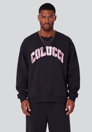 Mann, der eine Brille mit klarem Rahmen, geschichtete Goldketten, ein schwarzes Sweatshirt mit pinkem ""COLUCCI""-Text und passende schwarze Hose trägt, steht vor einem grauen Hintergrund.