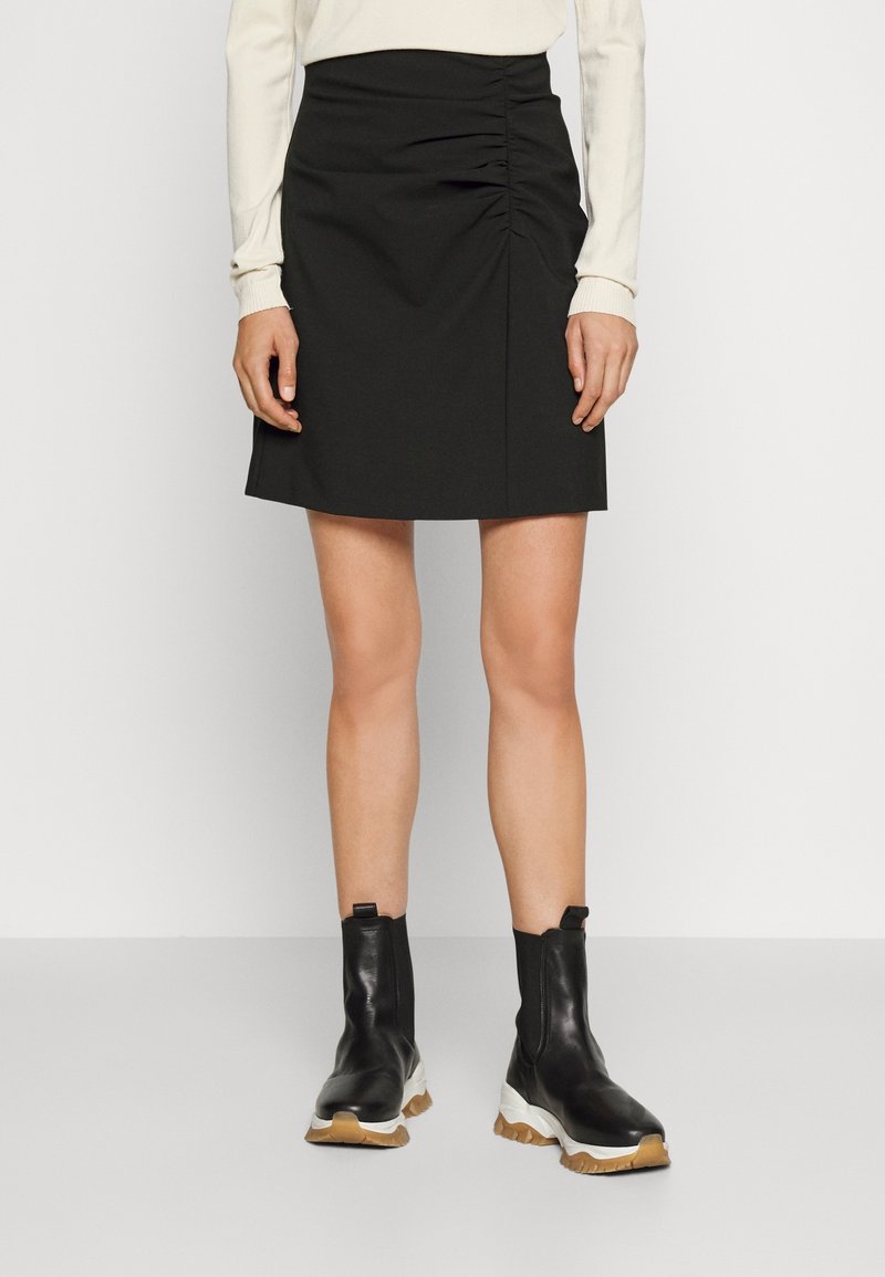 Černá přiléhavá sukně s nařaseným detailem na boku, vyrobená z hladké tkaniny. Spárována s černými kotníkovými botami s masivní podrážkou a texturovaným povrchem.