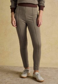 Brūnas un gai�šas krāsas houndstooth raksta legingi ar augstu vidukli un pieguļošu dizainu, kombinēti ar gaišiem sporta apaviem un fakturētu fonu.