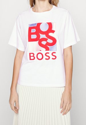 Personne portant un T-shirt blanc avec un motif graphique rouge, bleu et blanc portant la mention BOSS et une jupe plissée blanche.