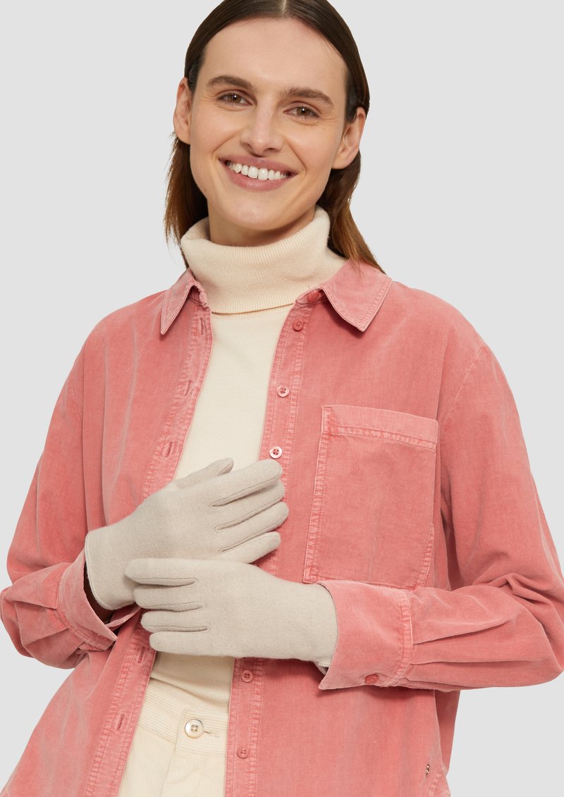 Beige handschoenen met een ontwerp met vijf vingers, weergegeven tegen een lichtroze corduroy shirt en een roomkleurige coltrui, die textuur en neutrale kleuren benadrukken.