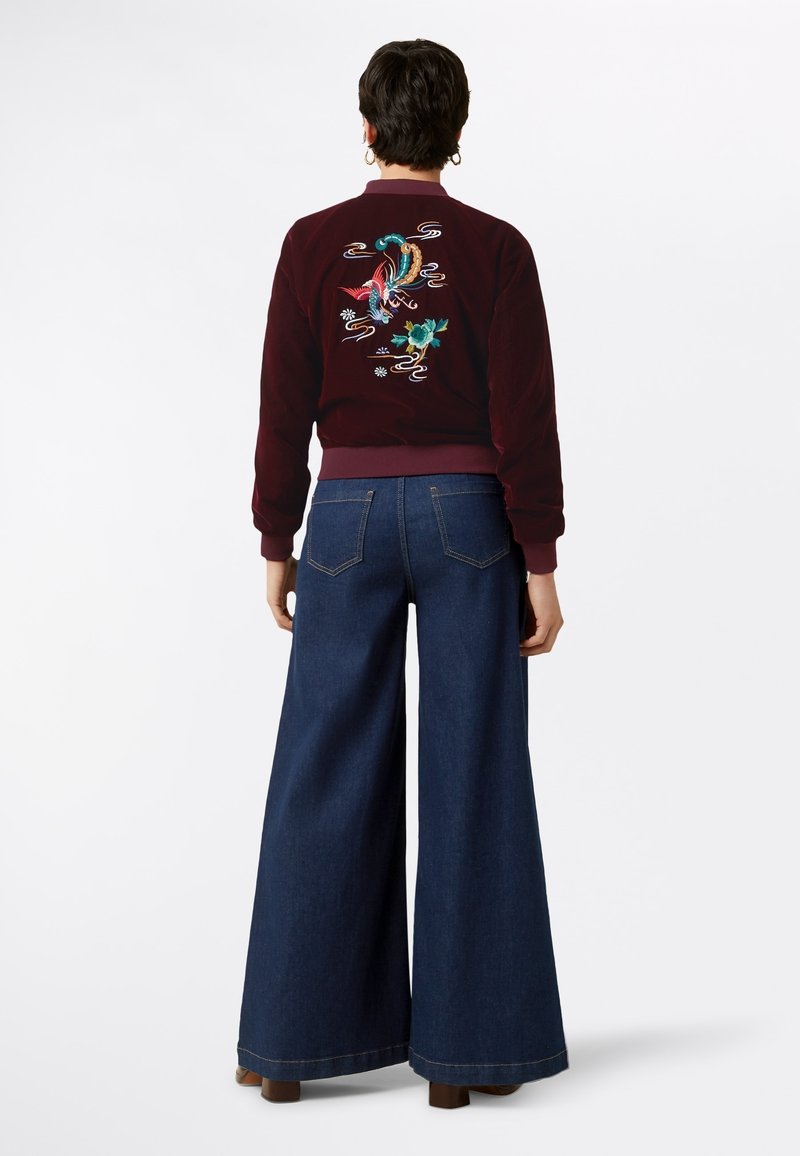 Personne portant une veste bomber grenat avec un phénix et des fleurs brodés colorés, associée à un jean bleu foncé à jambes larges et des chaussures marron.