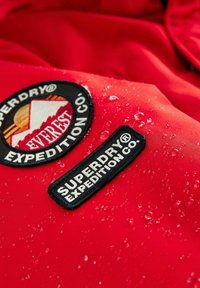 Červená nepremokavá tkanina má dva vyšívané nášivky: jednu s nápisom "Superdry Expedition Co." a druhú s dizajnom hory a nápisom "Everest."