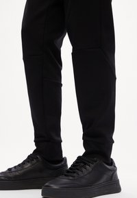 Benen in zwarte tapered pants en zwarte leren sneakers met veters op een witte achtergrond.