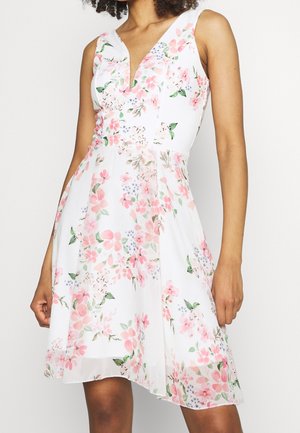 White sleeveless dress with pink and green floral pattern, fitted waist, and V-neckline, worn by a standing person with curly hair.