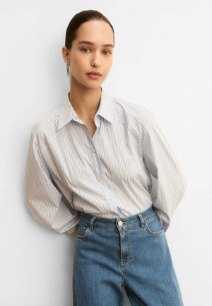 Donna che indossa una camicia a righe larga con maniche lunghe e bottoni, infilata in jeans blu a vita alta, in piedi contro un muro bianco semplice.