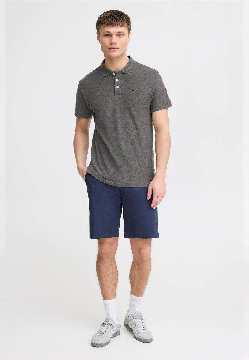 Gray polo shirt with three buttons, paired with navy shorts. Model wears gray sneakers and white socks, standing against a plain background.