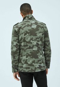 Viheret sävyissä oleva maastokuvioinen takki, jossa on korkea kaulus, joustava vyötärö ja napilliset olkataskut, nähtynä takaa.